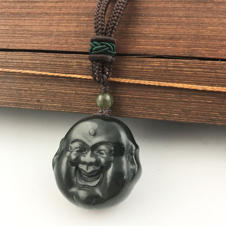 Circular black Buddha head pendant with a carved laughing face on a brown braided cord necklace with a green bead above.