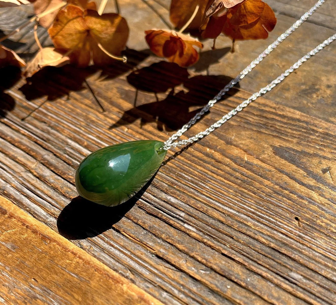 Green jade teardrop pendant on a silver chain resting on a sunlit wooden surface with autumn leaves.