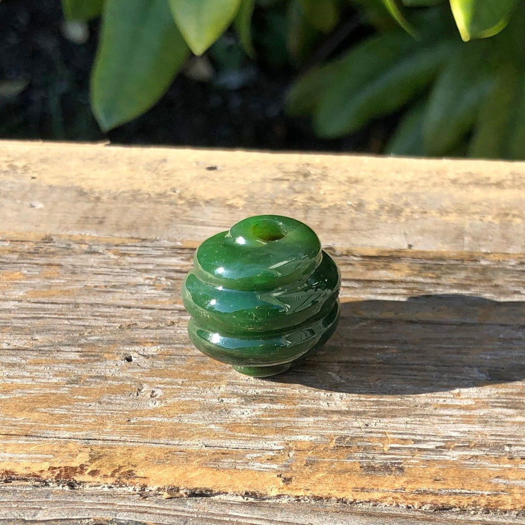 Green jade bead with layered spiral rings and a drilled hole sits on a wooden surface in sunlight