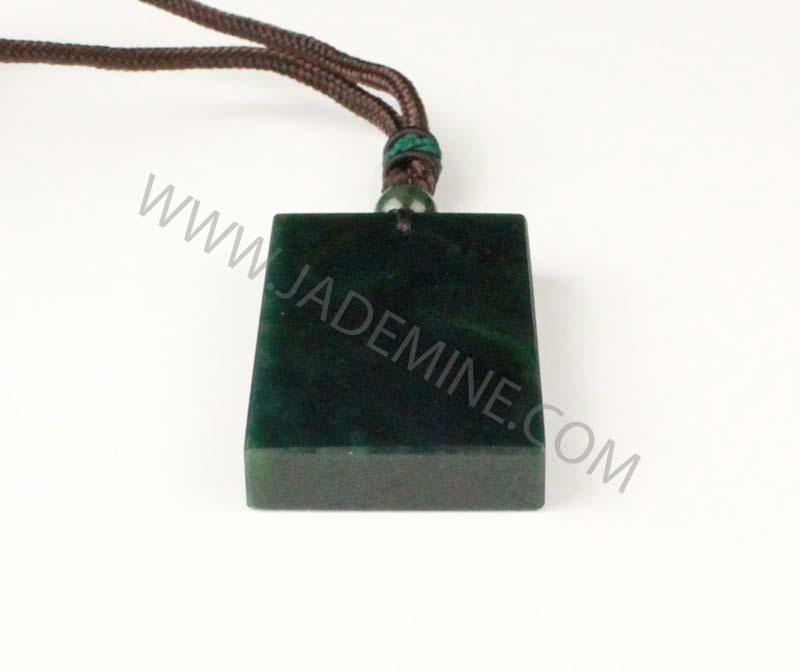 Dark green rectangular jade pendant with brown cord and bead resting on a white surface.