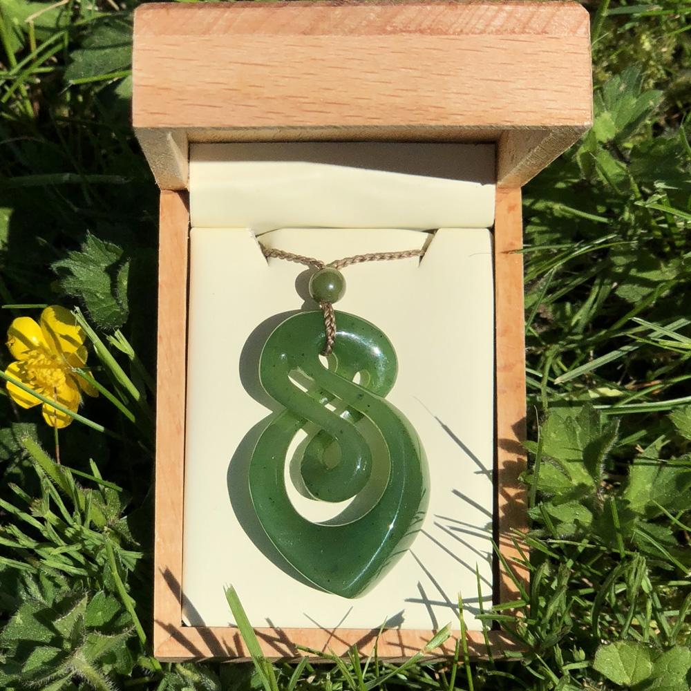Infinity Twist pendant in green jade inside an open wooden box placed on grass.