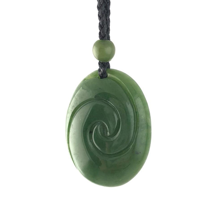 Close up of oval jade pendant with a spiral carving on a black braided cord and a green bead.