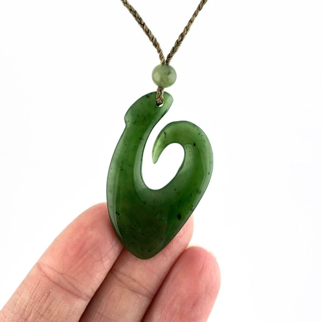 Canadian Jade fish hook pendant held between fingers on a braided tan cord with a bead above the hook.