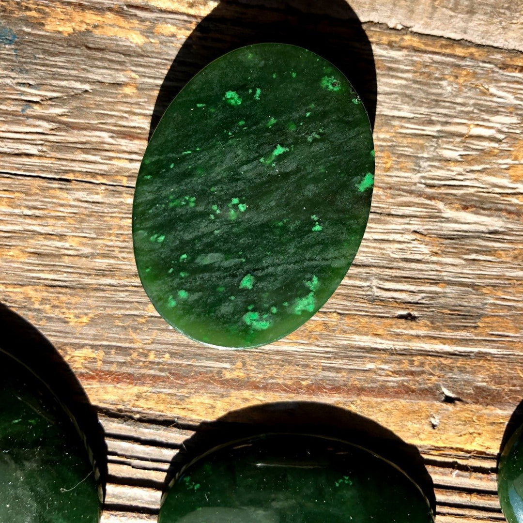 Single dark green Cassiar jade cabochon with subtle lighter green patches on a wood surface.