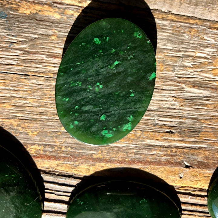 Single dark green Cassiar jade cabochon with subtle lighter green patches on a wood surface.