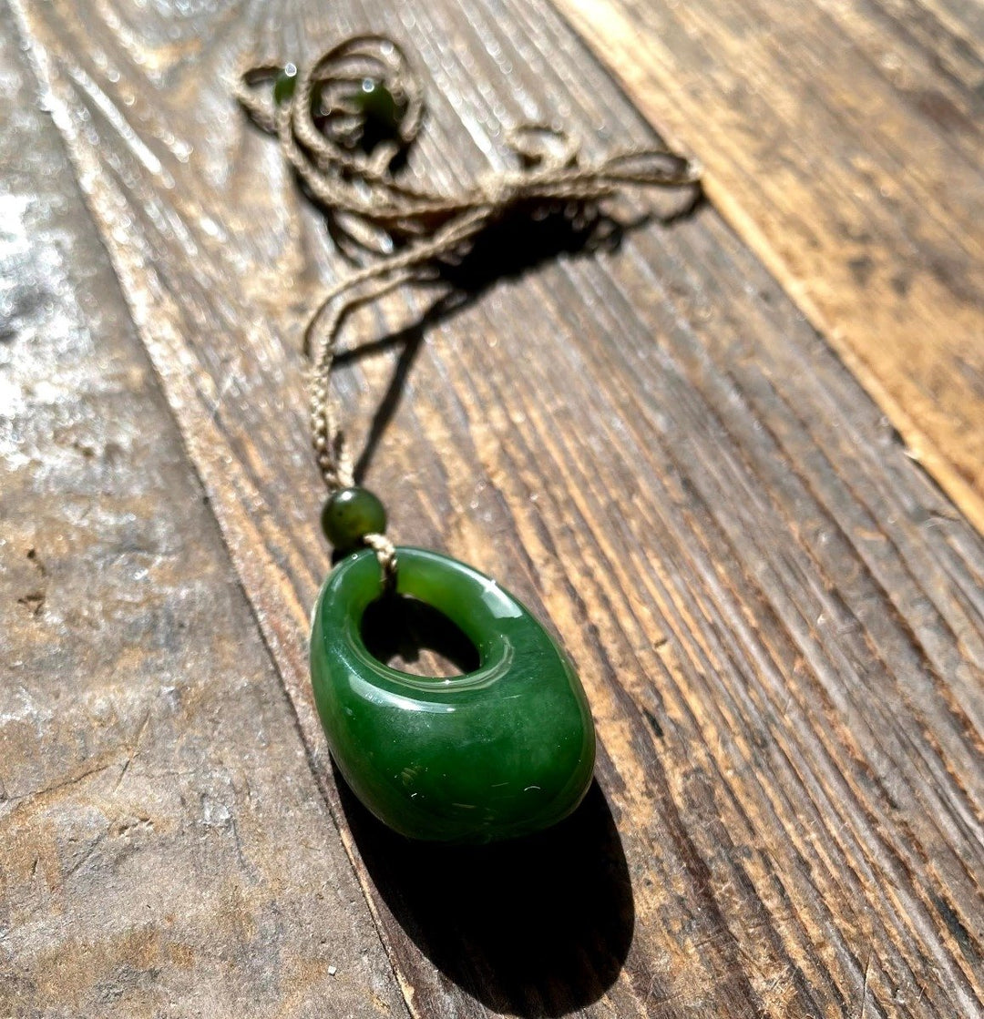 Close up of a jade donut pendant threaded on brown twine with green beads, on a weathered wooden surface.