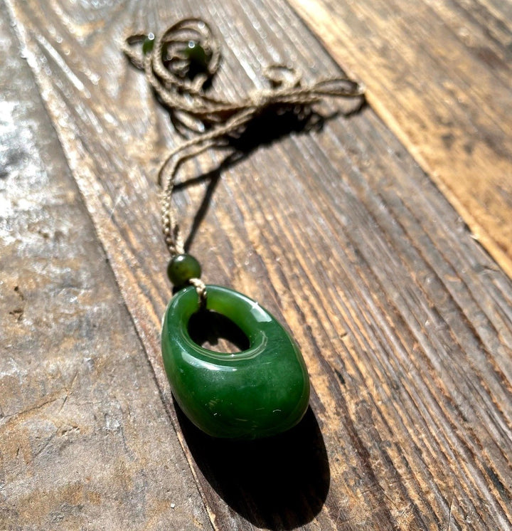 Close up of a jade donut pendant threaded on brown twine with green beads, on a weathered wooden surface.