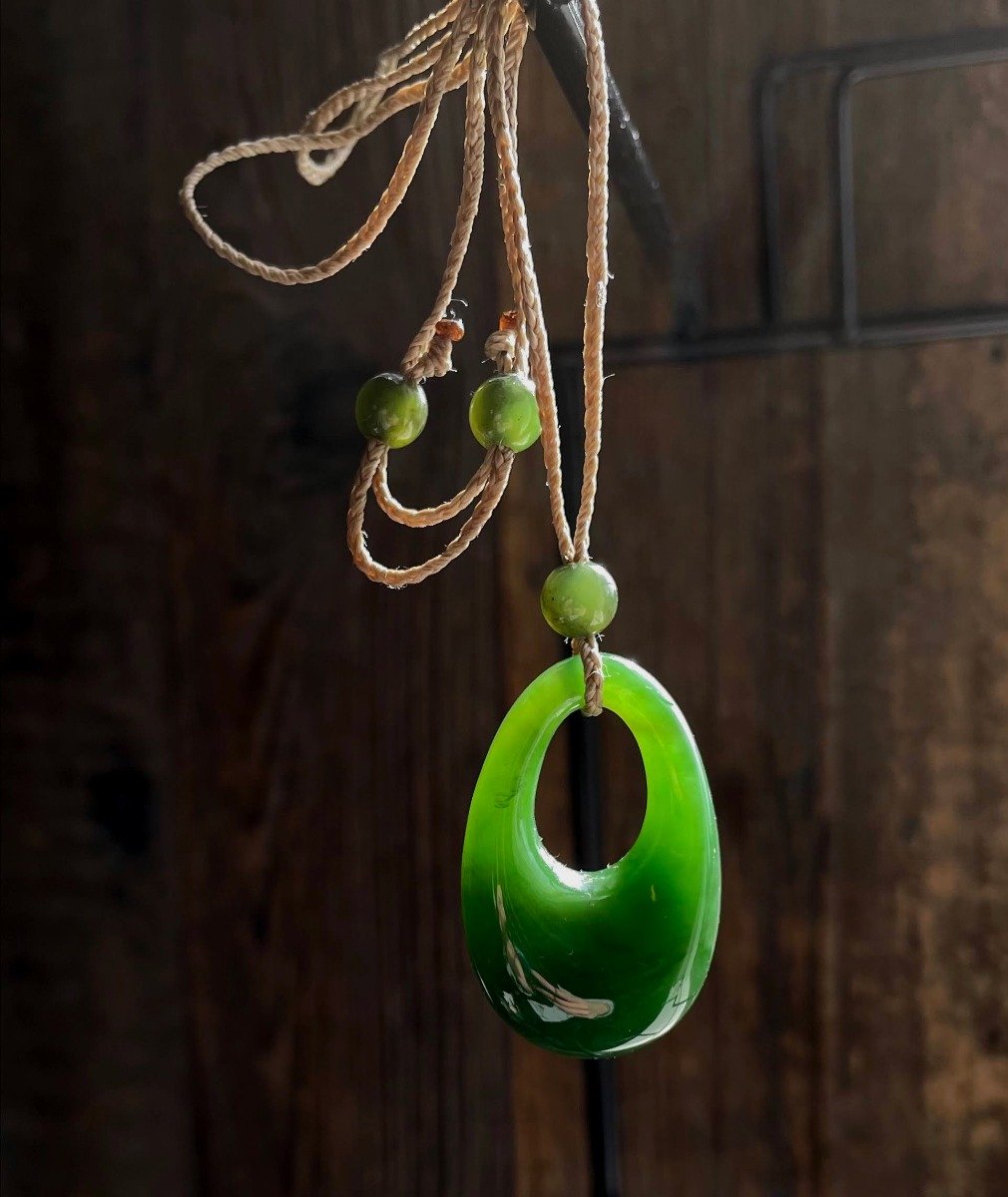 Bright green jade pendant shaped like a hollow oval on a beige cord with beads against dark wood background.