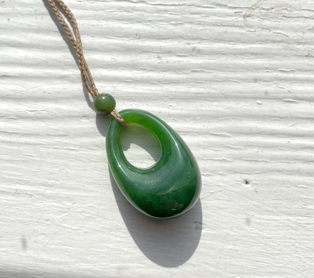 Green jade pendant with hole on a braided cord hanging against a pale textured wall
