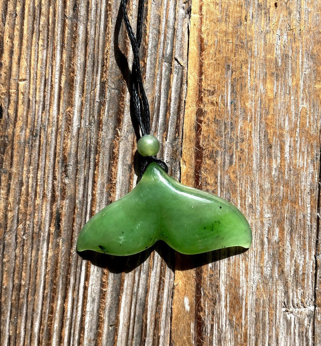 Jade whale tail pendant on a braided brown cord with a bead against sunlit wooden planks