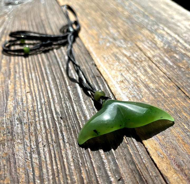 Green whale tail shaped jade pendant on a black cord with small green beads on weathered wood