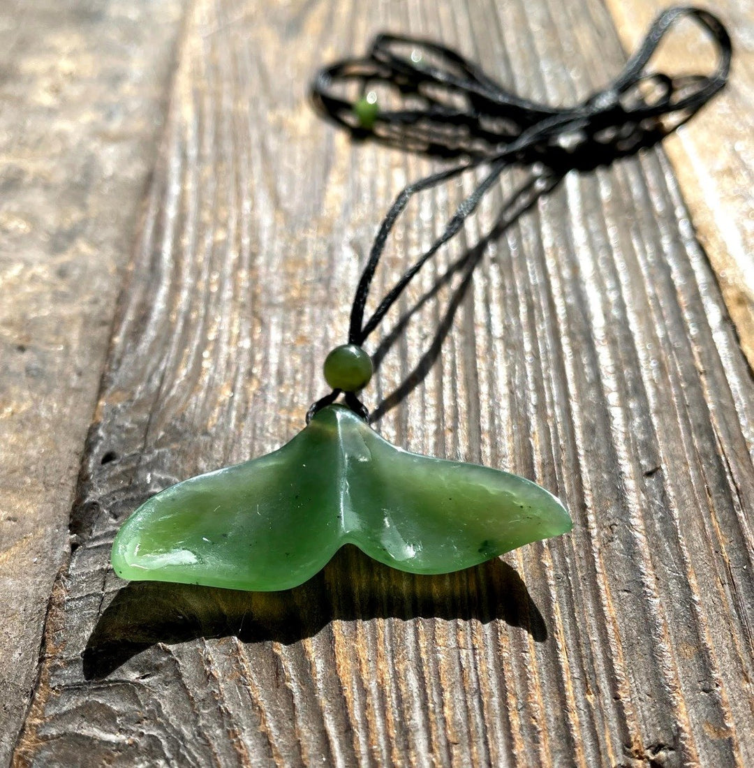 Green jade whale tail pendant resting on weathered wood with a black cord and bead.