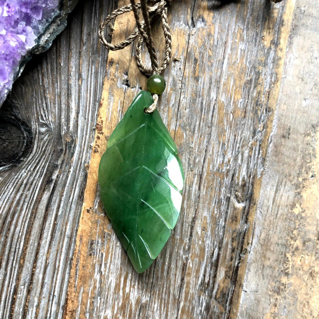 Green jade leaf pendant with a small bead on a braided cord against a rustic wooden background