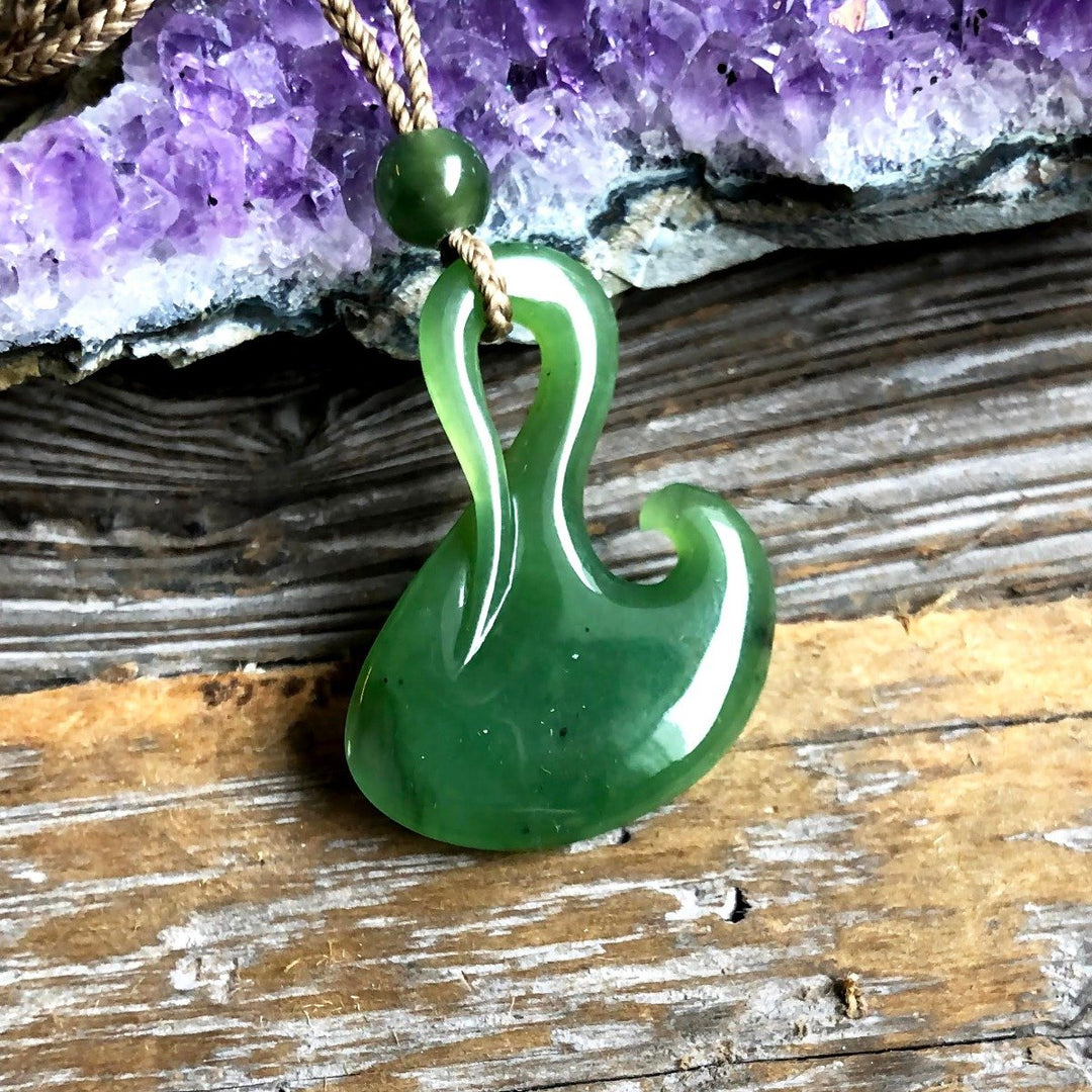 Green jade fish hook pendant with white edge detail on a braided cord against a purple crystal backdrop