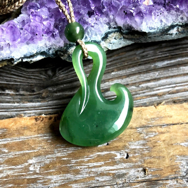 Green jade fish hook pendant with white edge detail on a braided cord against a purple crystal backdrop