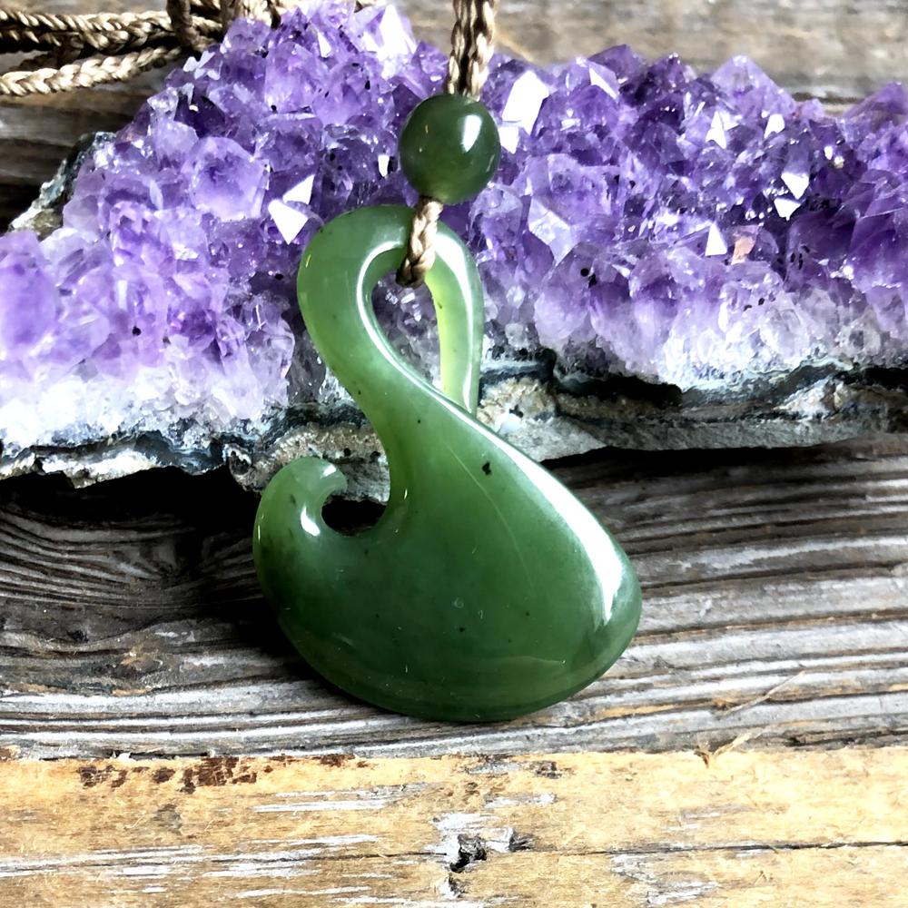 Green jade pendant shaped like a fish hook, with a bead and cord, against purple amethyst cluster.