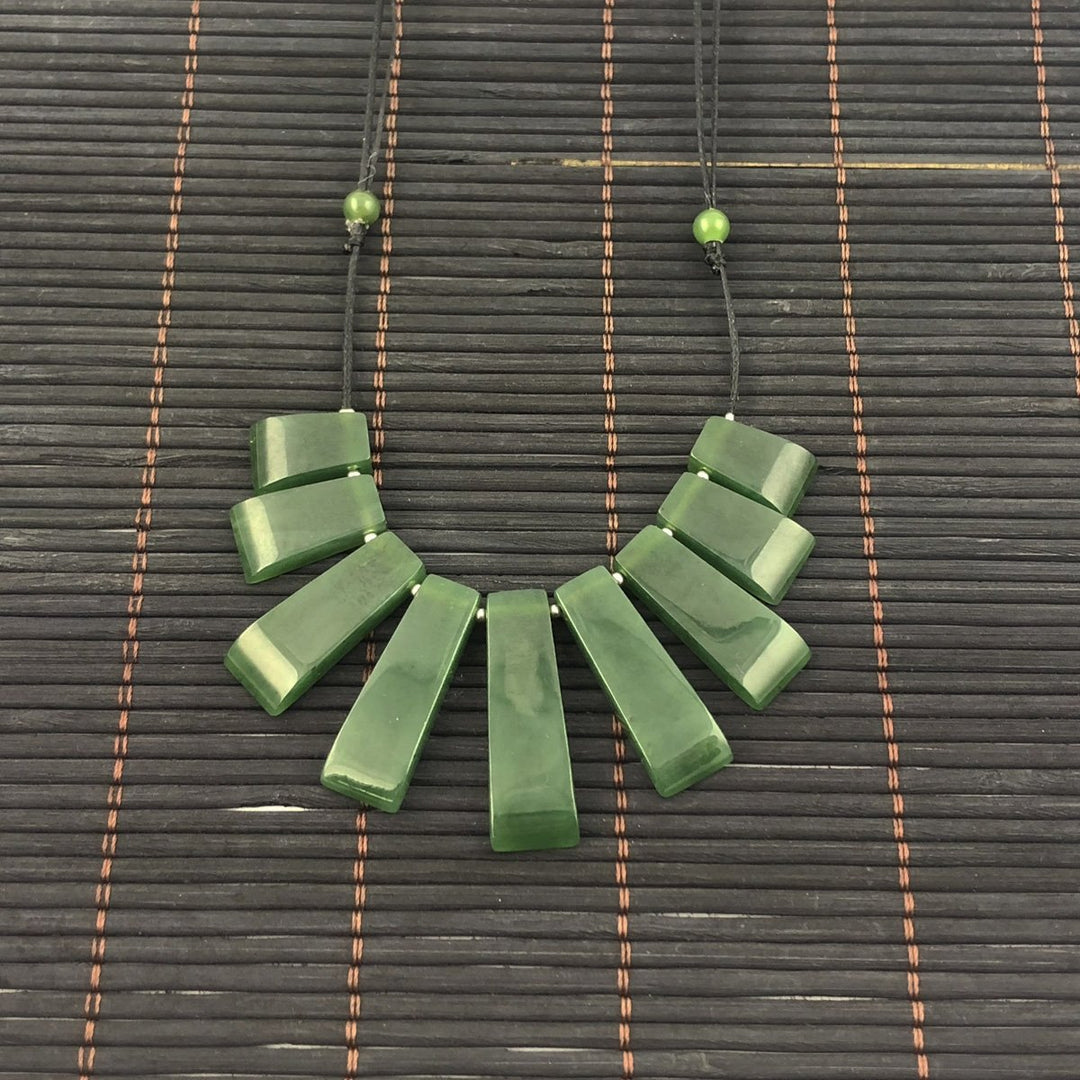 Jade necklace with long rectangular beads laid out in a fan shape on a black cord against a dark woven mat.