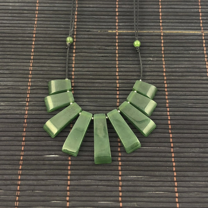 Jade necklace with long rectangular beads laid out in a fan shape on a black cord against a dark woven mat.