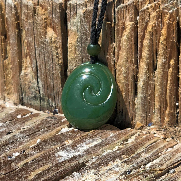 Green jade pendant with a circular spiral carving on a black braided cord against weathered wood background.
