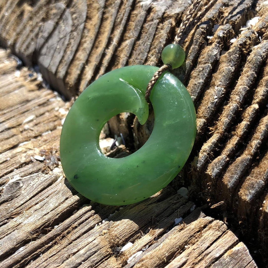 Green jade donut pendant on a braided cord with a small bead, shown on weathered wood.