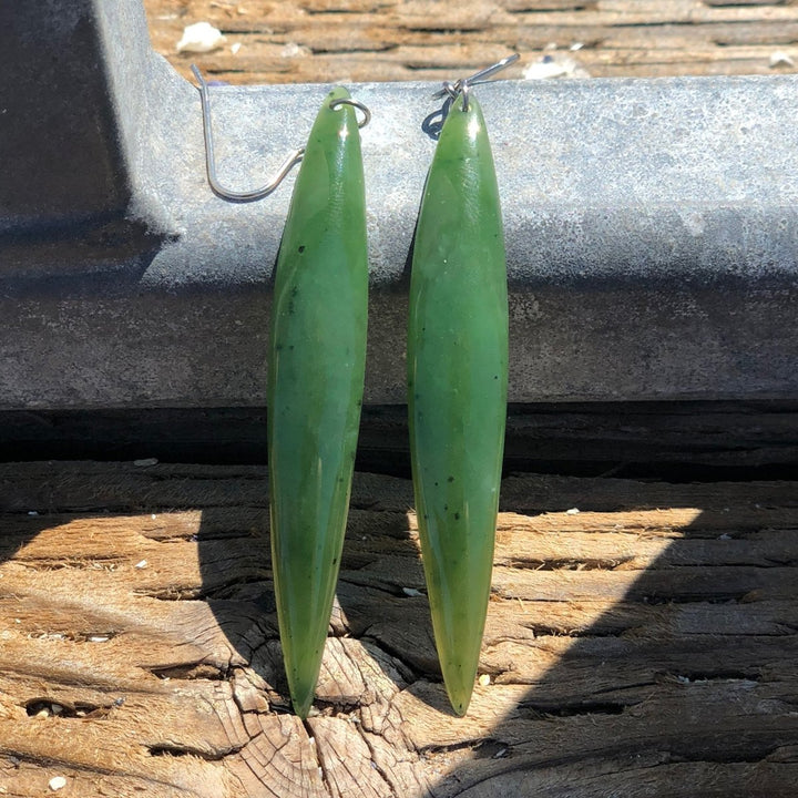 Close up of long green leaf shaped drop earrings with silver hooks resting on weathered wood.
