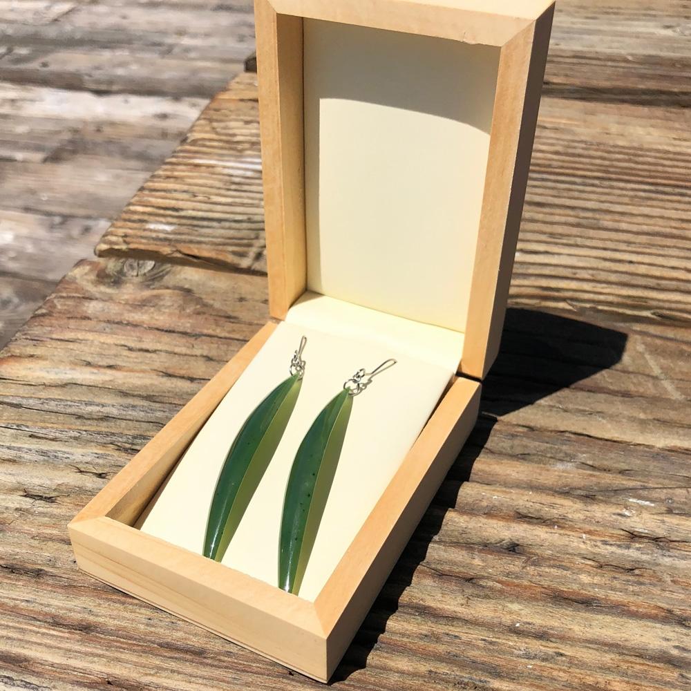 Long drop earrings with slim green leaf shaped pendants displayed in an open wooden box.
