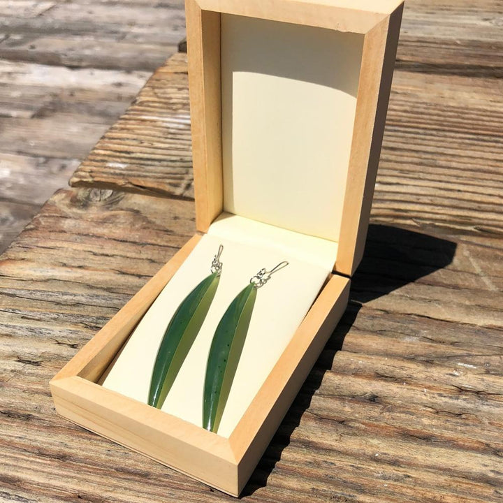 Long drop earrings with slim green leaf shaped pendants displayed in an open wooden box.