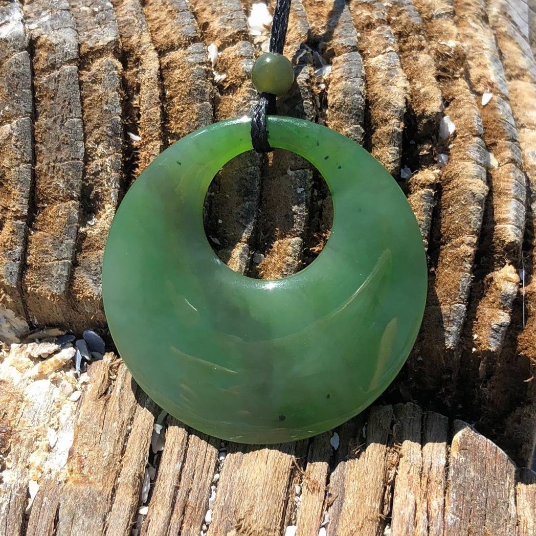 Green jade circle pendant with a central hole resting on a rustic log, threaded onto a black cord with a bead