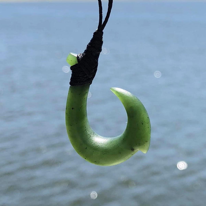 Light green jade hook pendant hanging from a black cord against a blue water background