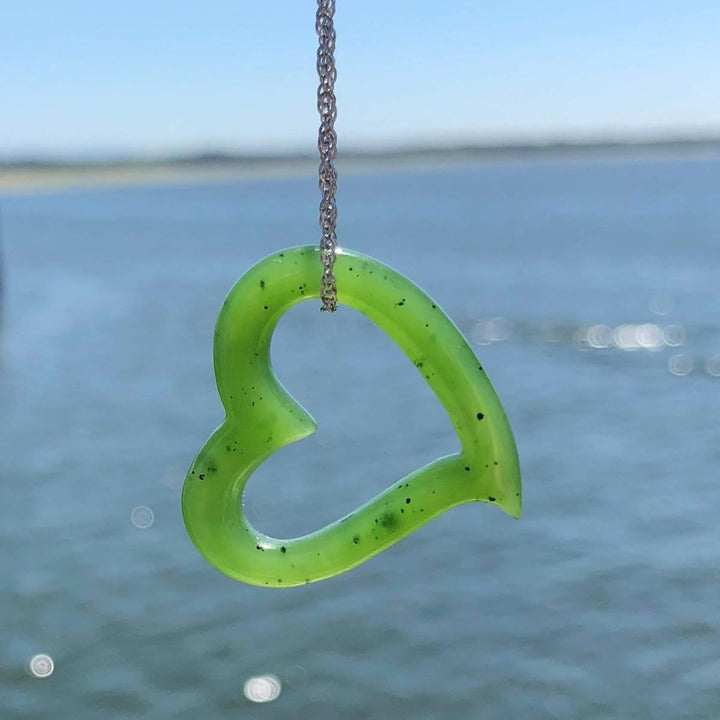 Translucent lime green heart shaped pendant hanging from a silver chain with a blurred ocean backdrop.