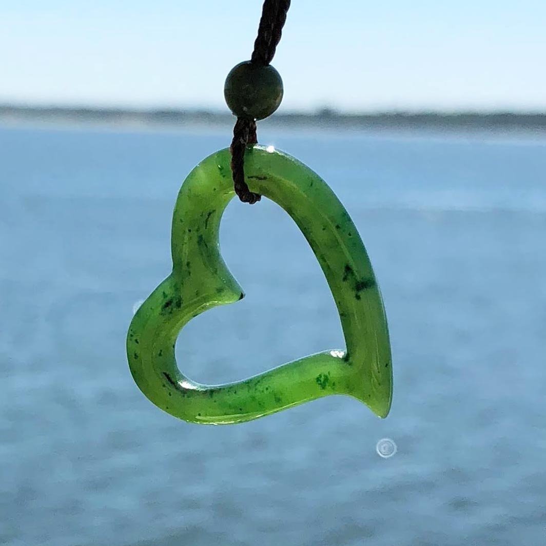 Green heart shaped pendant with hollow center on a dark brown cord against a blue sea backdrop