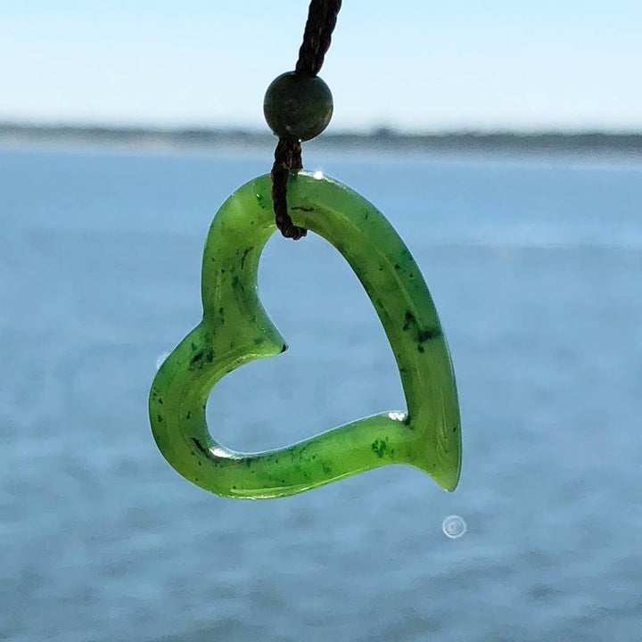 Green heart shaped pendant with hollow center on a dark brown cord against a blue sea backdrop