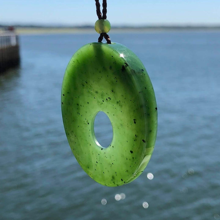 Green jade donut pendant hanging from braided brown cord over a blue water backdrop