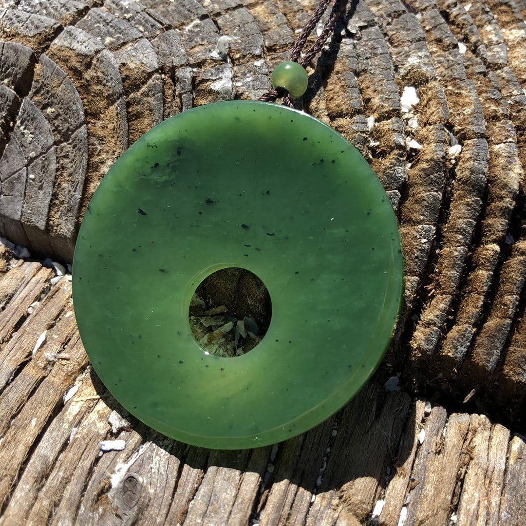 Green jade donut pendant with a small bead on a brown cord resting on weathered driftwood