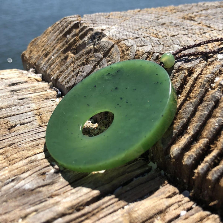 Green jade donut pendant on a brown cord laid on textured wood with water in the background