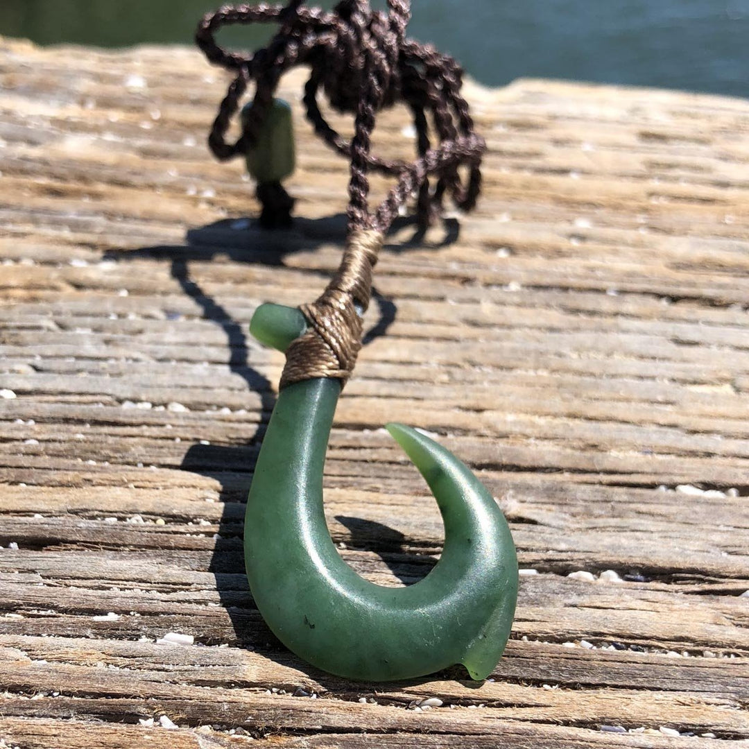 Matte green jade fish hook pendant with a brown braided cord on a weathered wooden surface.