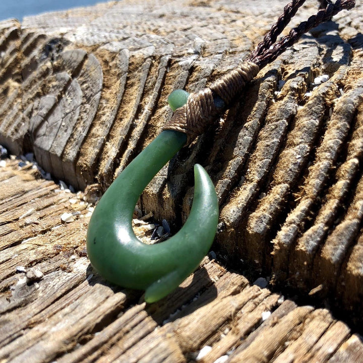 Green jade fish hook pendant with a brown cord wrapped around the rope, set against textured driftwood near the water.