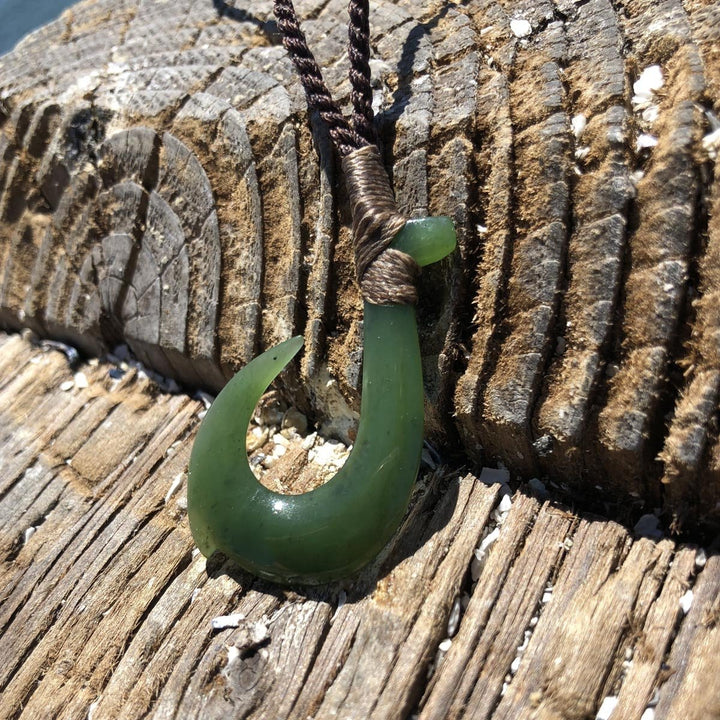 Jade fish hook pendant wrapped with brown cord against weathered driftwood.