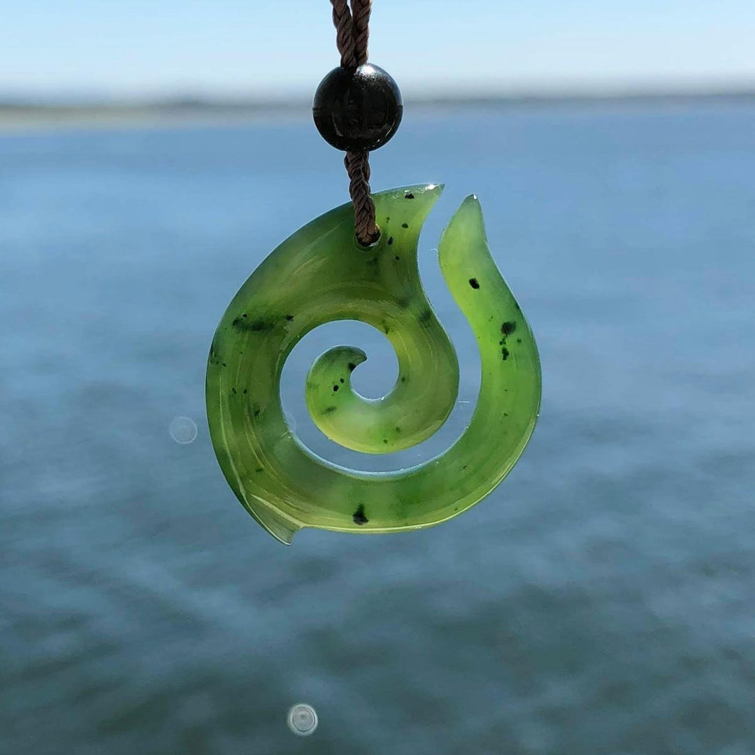 Green jade spiral pendant on brown cord with black bead over ocean backdrop