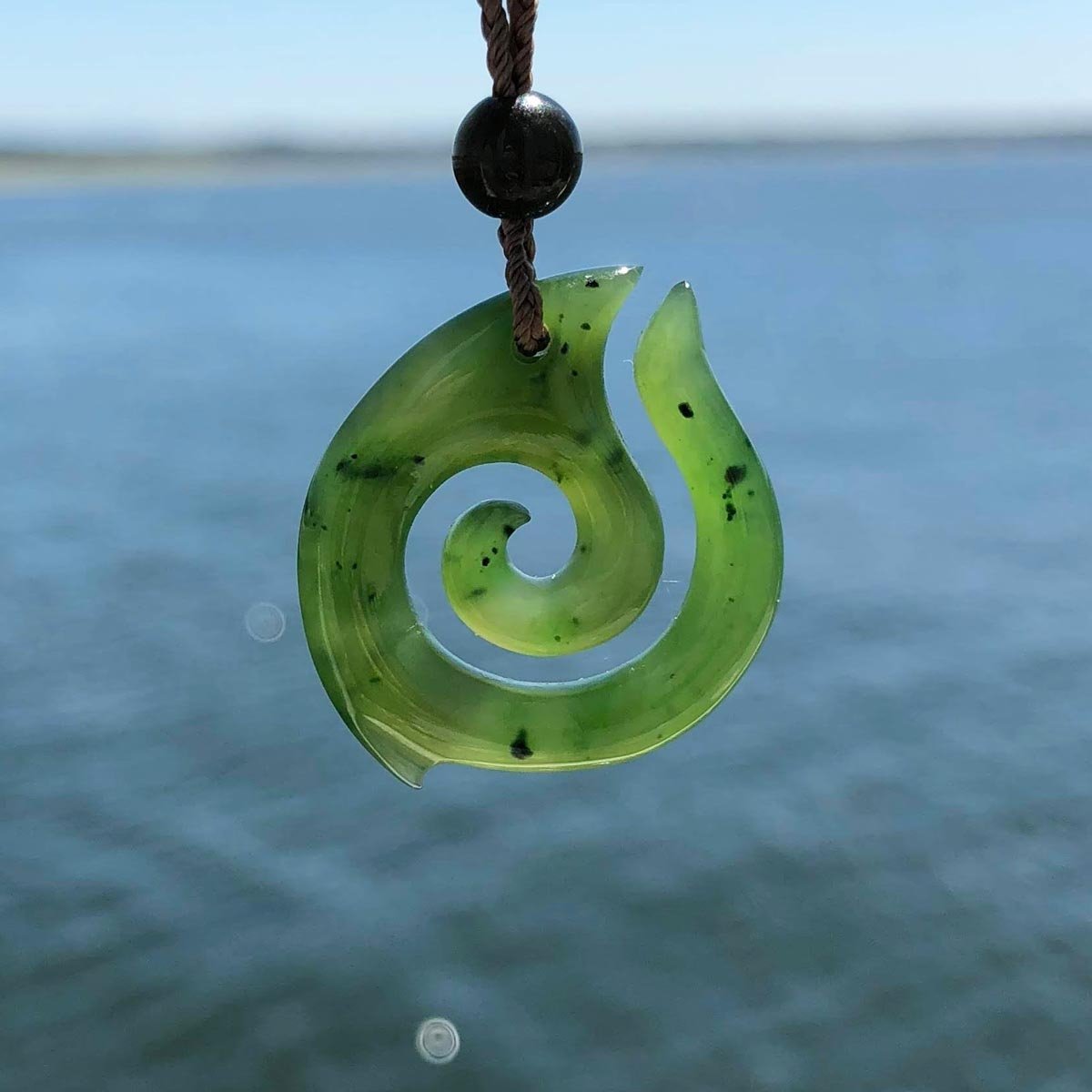 Green jade spiral pendant on brown cord with black bead over ocean backdrop