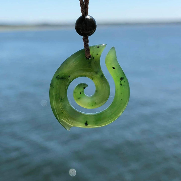 Green jade spiral pendant on brown cord with black bead over ocean backdrop