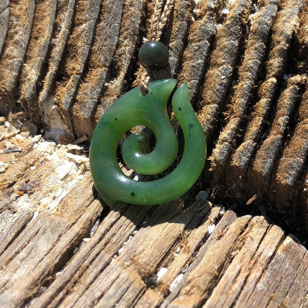 Green jade spiral pendant on a braided cord with a round bead, set against weathered wood.