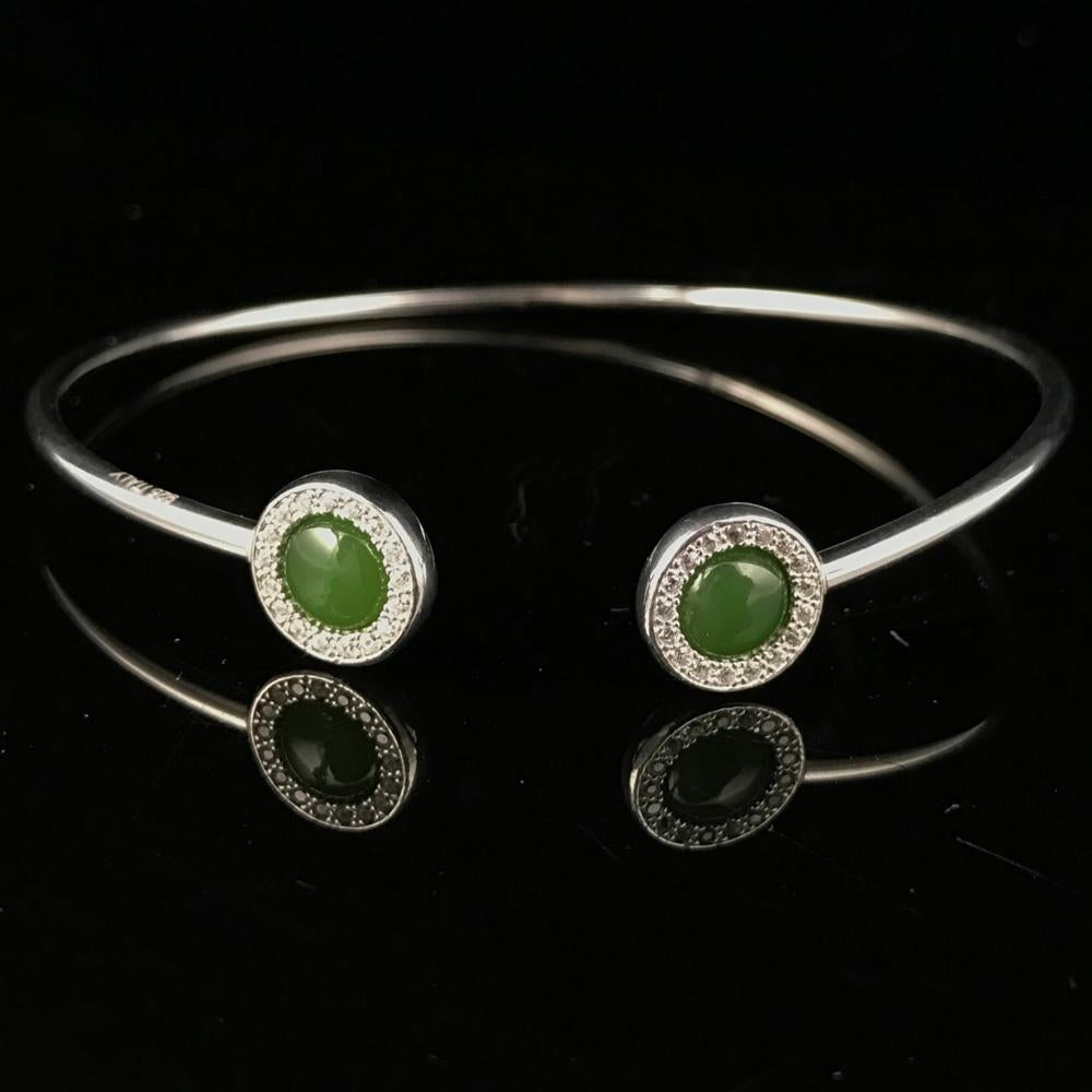Sterling silver open cuff bracelet with two green stones at the ends and pavé halos on a reflective black surface.