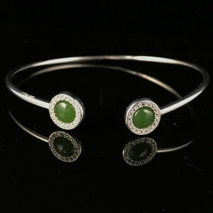 Sterling silver open cuff bracelet with two green stones at the ends and pavé halos on a reflective black surface.