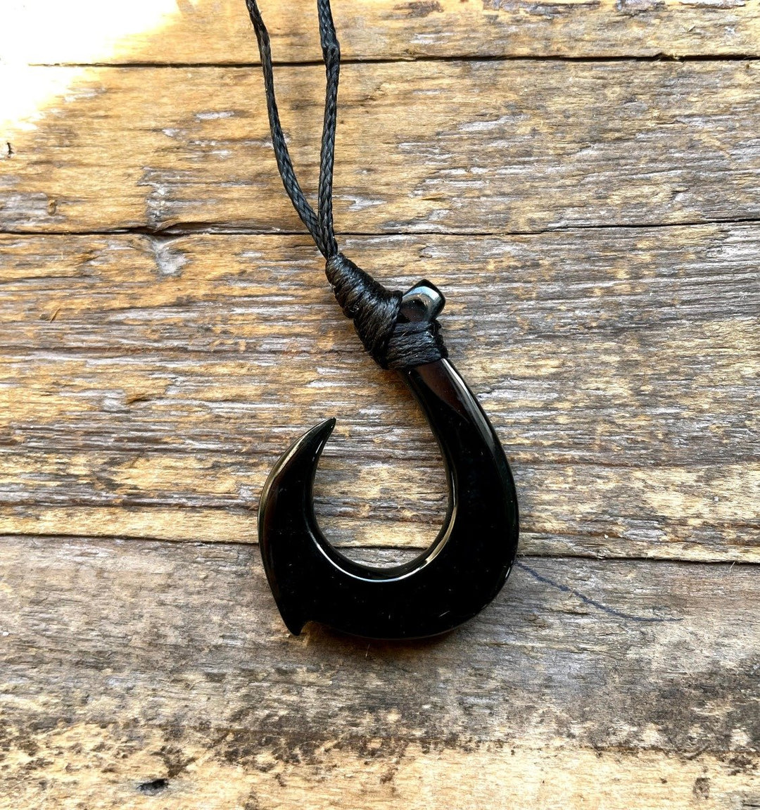 Jade fish hook pendant on braided black cord against a weathered wooden surface