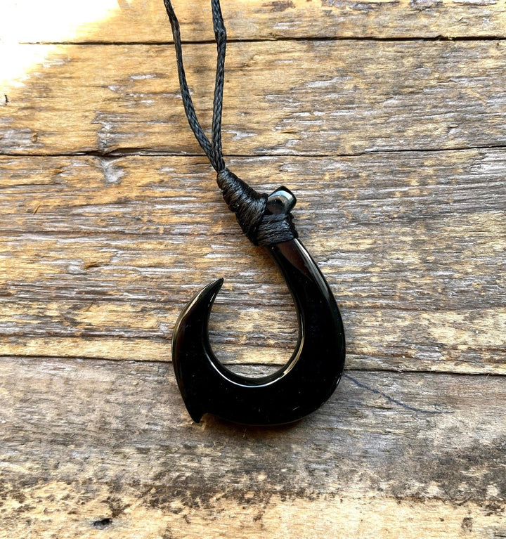 Jade fish hook pendant on braided black cord against a weathered wooden surface