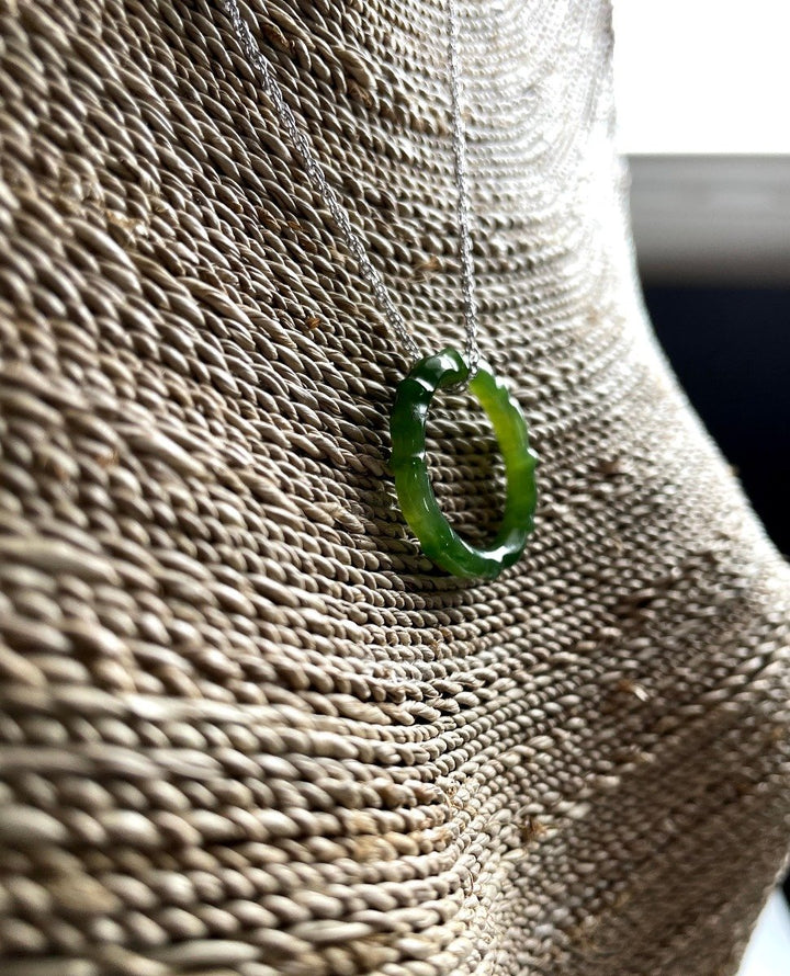 Green jade circular pendant on a silver chain resting against a woven bag background