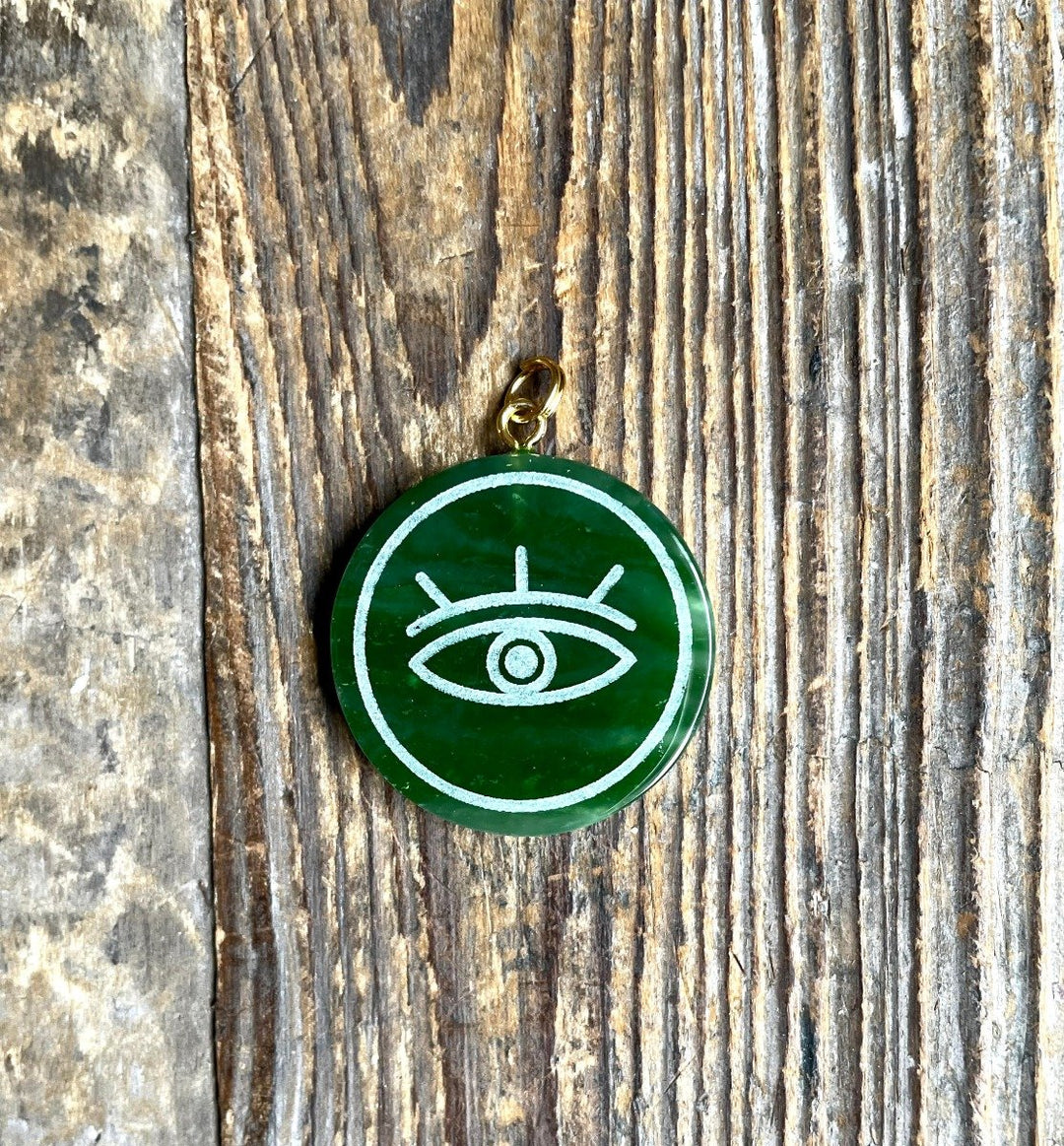 Green circular jade evil eye pendant with white engraving and gold bail on rustic wood