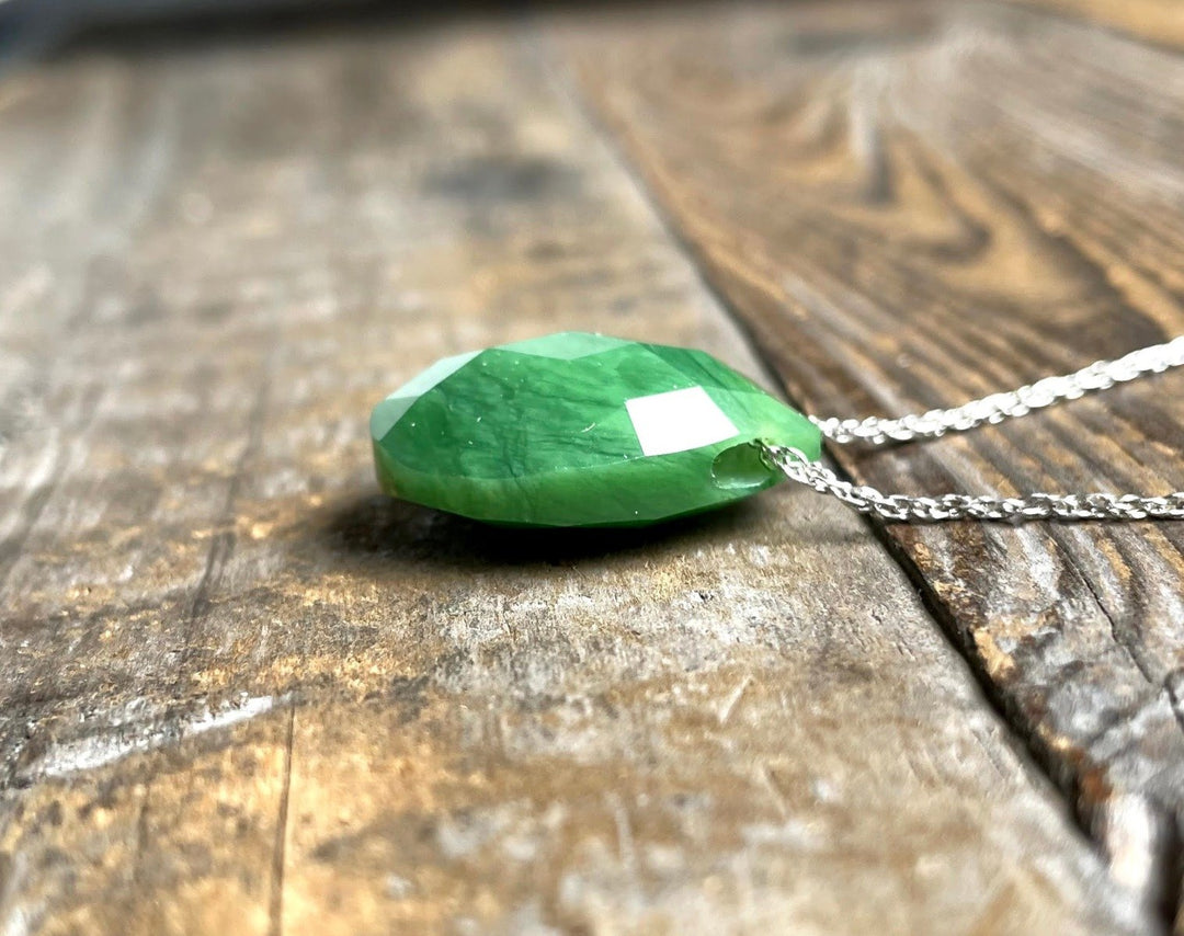 Faceted emerald green teardrop pendant on a silver chain resting on rustic wooden surface