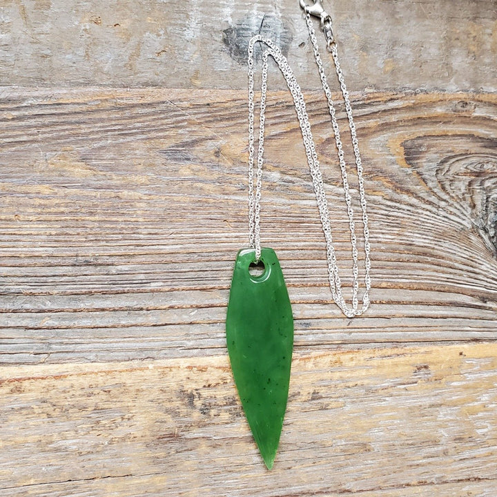 Green jade drop pendant with a top hole on a silver chain resting on a rustic wooden surface.